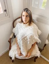 Poncho de punto color marfil, confeccionado en lana merino, con cuello alto, diseño de rombos texturizados con bodoques y flecos voluminosos en los bordes laterales.