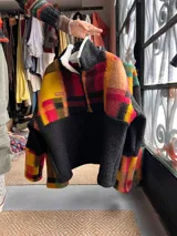 Buzo canguro de corderito estampado a cuadros con predominio de rojo, amarillo y negro, con cuello alto y cierre frontal parcial.