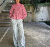 Blusa con estampado de cuadros rojos y blancos, cuello alto y cuerpo fruncido tipo nido de abeja. Presenta mangas largas abullonadas con puños elásticos y un volante en el bajo estilo peplum.