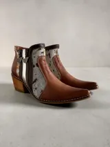 Botas cortas de cuero en tonos marrón y tostado, con paneles de cuero vacuno con estampado animal print tipo ponchada y detalles pintados a mano en dorado. Presentan punta fina, taco cuadrado de madera, costuras visibles a contratono y doble cierre lateral con tiradores metálicos personalizados.