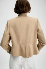 Blazer entallado de sarga color beige con cuello subido y escote en pico. Presenta manga larga con hombreras, detalle de botones metálicos decorativos en el frente y cierre frontal con gancho metálico. Incluye falsos bolsillos de vivo y abertura en la espalda.