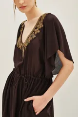Vestido corto de color negro con escote en V profundo, adornado con bordados dorados y pedrería. Presenta mangas cortas tipo mariposa, cintura ajustable con cordón y bolsillos laterales.