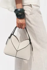 Bolso tipo mini shopper confeccionado en material trenzado color beige, con detalles en cuero sintético marrón. Presenta una estructura rígida con forma de V en la parte superior, asas de mano fijas y correa bandolera extraíble. Incluye una cartera interior con cierre.