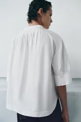 Camisa blanca de algodón con cuello redondo y manga corta abullonada. Presenta un canesú con pliegues verticales en la parte delantera y cierre frontal con botones ocultos. El bajo es redondeado.