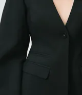 Blazer de sastrería negro de corte entallado, sin cuello, con escote en V profundo, manga larga y cierre frontal de un botón. Cuenta con bolsillos frontales con tapa, recortes estructurados y tira trasera con hebilla.