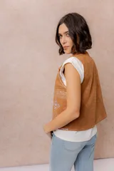 Chaleco corto de efecto gamuza color tostado, con detalles bordados en contraste en los bordes y en la parte inferior. Presenta escote en V y diseño abierto al frente.