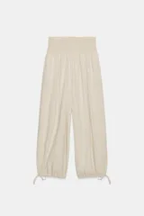 Pantalón ancho color beige claro, de tiro alto con cintura fruncida elástica. Las piernas son anchas y terminan en puños ajustables con cordones.