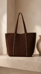 Cartera tipo tote bag de símil gamuza color marrón oscuro, de diseño amplio con detalles de tachas metálicas en los bordes y correas de hombro.