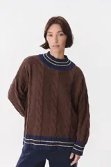 Buzo de punto con diseño de trenzas, color marrón con detalles en azul marino y líneas amarillas en cuello, puños y ruedo. Presenta un corte oversized con hombros caídos.