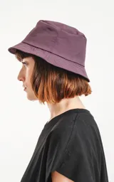 Piluso de estilo bucket hat en color violeta oscuro, con costuras circulares en el ala y parche cuadrado frontal con logo de tijeras.