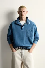 Camisa polo de corte relajado confeccionada en denim ligero de algodón con efecto lavado. Presenta cuello subido, cierre frontal con botonadura oculta por solapa, manga larga, bolsillo de plastrón en el pecho y bajo ajustable con cordón.