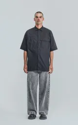 Camisa de manga corta en popelina de algodón, con diseño oversize y cierre frontal de botones. Presenta dos bolsillos delanteros, uno con solapa y otro con ojal y botón.