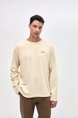 Remera de manga larga color beige, confeccionada en algodón lavado con textura suave. Presenta cuello redondo con detalle de costura en V, corte clásico y estampado pequeño en el pecho con la palabra Uniform.
