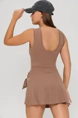Vestido corto deportivo color habano, con diseño sin mangas, cuello redondo y detalle de falda envolvente con lazo lateral. Incluye logo pequeño bordado en el pecho.