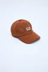 Gorra de pana color marrón caramelo con visera curva y parche rectangular blanco con texto negro en el frente.