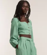Blusa cropped verde menta de sastrería, confeccionada en viscosa y poliéster, con escote cuadrado y mangas largas abullonadas con puños elásticos. Presenta cierre frontal con botones matizados.