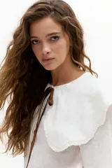 Camisa blanca de lino con cuello Peter Pan grande y ondulado, mangas largas y cierre frontal con botones de nácar. Incluye lazo decorativo de terciopelo marrón en el cuello.