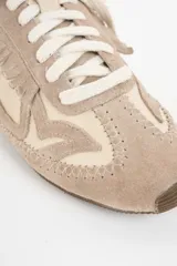 Zapatilla de cuero legítimo en tonos beige y crema, con diseño de flecos en el lateral y costuras decorativas visibles en el contorno. Presenta cordones al tono y suela plana.