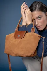 Cartera tipo bolso de mano confeccionada en gamuza color suela, con solapa de cuero liso y cierre metálico central. Incluye manijas cortas y correa larga ajustable y extraíble.