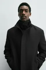 Abrigo negro de corte recto con cuello subido y doble cuello desmontable mediante cremallera interior. Cuenta con manga larga, bolsillos de vivo en la cadera, bolsillo interior, abertura central en la espalda y cierre frontal de cremallera oculta por solapa con botones a presión.