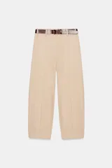 Pantalón de tiro bajo color beige, de corte globo, con bolsillos delanteros, costuras frontales marcadas y cierre frontal con cremallera, botón interior y gancho. Incluye cinturón de cuero marrón con hebilla metálica.