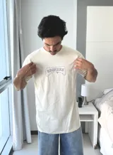 Remera de manga corta color beige jaspeado, con un estampado gráfico en la espalda que incluye el texto "TAS LOCO" y "SUNNY WITH A CHANCE OF STOKE" con un diseño de una persona en el agua con una ballena.