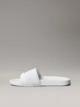 Ojotas tipo slide Calvin Klein Jeans, color blanco, con banda superior de lona de algodón que presenta el monograma de la marca en color negro. Posee suela de EVA con plantilla ergonómica moldeada.
