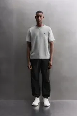 Camiseta relaxed fit de algodón, color gris melange con mangas en un tono de gris más claro. Tiene cuello redondo, manga corta, bordado frontal y texto estampado en la espalda a contraste. Incluye un parche con logo en el bajo.