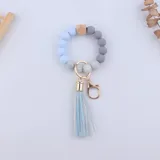 Llavero pulsera con cuentas redondas de silicona en tonos celeste, gris y blanco, con un cubo de madera natural y borla decorativa celeste. Incluye gancho metálico dorado.