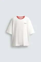 Camiseta de corte relajado, color blanco, con cuello redondo en contraste rojo y manga corta. Presenta un pequeño estampado en el pecho con texto cursivo y el número "99" en color blanco tenue.