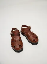 Sandalias tipo romanas de cuero vacuno color marrón, con diseño de tiras entrelazadas, cierre con hebilla metálica lateral y suela de goma dentada con plataforma.