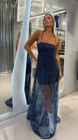 Vestido largo de fiesta color azul marino con diseño de brillos en patrón de hojas. Presenta un corte tipo strapless con breteles finos, una falda corta interior y una sobrecapa de tul transparente con tajo lateral.