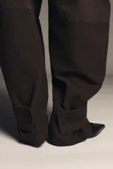 Pantalón cargo de algodón color marrón oscuro, tiro medio, con pliegues frontales, bolsillos delanteros ocultos y bolsillos de plastrón con solapa y botones a presión en las piernas. El bajo del pantalón tiene una trabilla con botón y se muestra doblado hacia afuera.