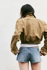 Trench corto de cuello solapa y manga larga, con diseño cruzado y cierre frontal con botones. Presenta bajo acabado en elástico y trabillas en los hombros.