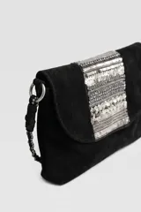 Cartera bandolera de cuero negro con solapa, decorada con apliques bordados de lentejuelas y abalorios plateados en el centro. Incluye una correa larga de hombro estilo cadena trenzada.