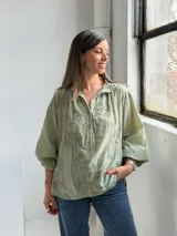 Camisa verde claro de corte oversize, con mangas tres cuartos abullonadas, escote en V con cuello camisero y cierre frontal parcial de botones. Cuenta con delicados bordados florales tono sobre tono distribuidos por toda la prenda.