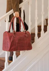 Bolso de viaje tipo duffel confeccionado en cuero color bordó, con detalles de costuras decorativas verticales y herrajes metálicos dorados. Cuenta con doble asa de mano y cierre superior.