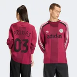 Suéter de punto Adidas en color bordó, inspirado en el estilo de las camisetas de fútbol retro. Presenta cuello redondo, mangas largas con las tres tiras características de la marca en blanco y detalles perforados en la trama. Incluye el logo del Trifolio y un escudo circular bordados en el pecho, junto con el texto "adidas" en contraste.