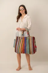 Bolso de hombro tipo tote grande, confeccionado con tejido de yute o fibras naturales con diseño de rayas verticales multicolor en tonos tierra, azul, verde, amarillo, rojo y naranja, con asas dobles fijas.