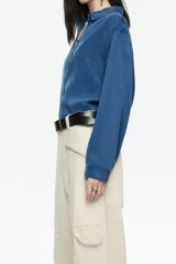 Camisa de manga larga color gris claro con efecto lavado, corte recto y botones frontales marrones. Presenta cuello camisero y pespuntes a contraste.