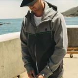 Campera canguro Maui and Sons de felpa con cierre completo y capucha ajustable con cordón. Presenta un diseño bicolor en tonos gris claro y gris oscuro, separado por una franja horizontal con patrón de olas y detalles en turquesa. Cuenta con bolsillos laterales tipo ojal y puños y ruedo de rib elástico.