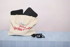 Bolso tipo tote de tela color crema con asas largas. Estampado frontal con la frase "NACIDA PARA FLOTAR" en azul y "SRTAPEEL.COM" en rojo.