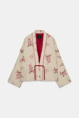 Chaqueta acolchada color beige con bordados florales en tonos rosados y rojos. Tiene escote en pico, manga larga y forro interior rojo. El cierre frontal es con alamar y tiene ribetes rojos.
