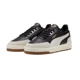 Championes Puma modelo Shuffle Downtown OG, de diseño urbano con base blanca, detalles en negro y suela de goma color caramelo. Presentan refuerzos en gamuza gris en la puntera y el talón, con el logo de la marca en dorado.
