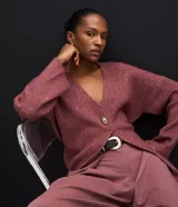 Cardigan de tricot en color borravino, con textura acanalada, escote en V profundo y cierre frontal con un único botón metalizado. Presenta mangas largas y un diseño alargado con amarre ajustable en la espalda.
