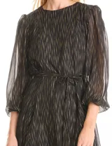 Vestido corto de gasa negra con estampado de rayas verticales doradas y plateadas. Tiene mangas largas semitransparentes con puños elastizados, cuello redondo, cintura con lazo ajustable y falda evasé con vuelo. La espalda presenta una abertura tipo lágrima con botón en la nuca.