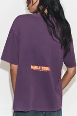 Remera oversize de algodón con efecto lavado en color violeta. Presenta cuello redondo, manga corta y una estampa frontal colorida con una ilustración artística de Manuja Waldia.