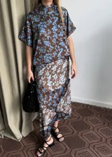 Blusa de seda con estampado floral en tonos marrón y celeste, con cuello alto, manga corta japonesa y cierre cruzado con botones negros visibles en diagonal.