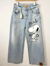 Jean de corte wide leg en denim azul claro con lavado desgastado. Presenta una ilustración pintada a mano de Snoopy en la pierna derecha, acompañada por la palabra 'Real', una corona y una estrella. Incluye etiqueta de cuero en la cintura y en el ruedo.