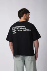 Remera de corte boxy y fit cuadrado, confeccionada en algodón grueso de alta calidad. Presenta un diseño minimalista en el frente con un pequeño logo circular y una estampa de gran tamaño en la espalda con la frase 'Designed in South America, Worldwide Clothes'.